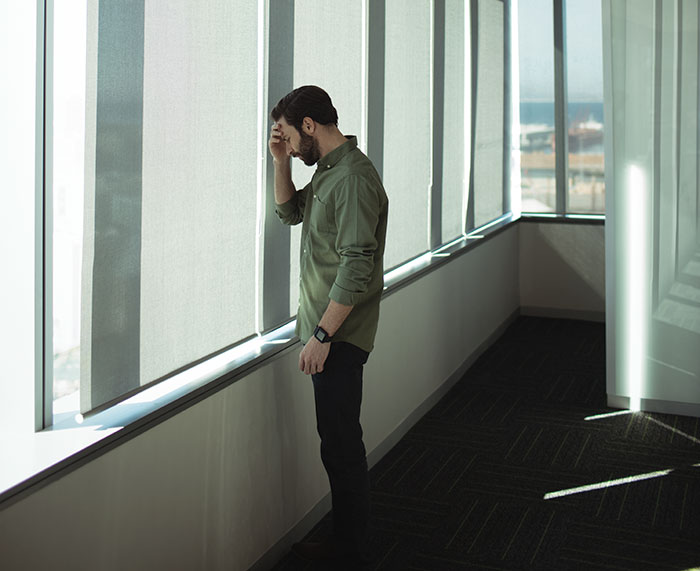 Man in green shirt looking out office window appearing confused by social cues on a bright day indoors