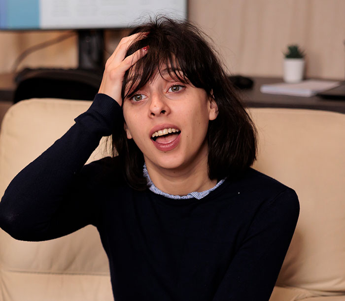 Young woman looking confused by social cues, sitting on a beige couch in a casual indoor setting.