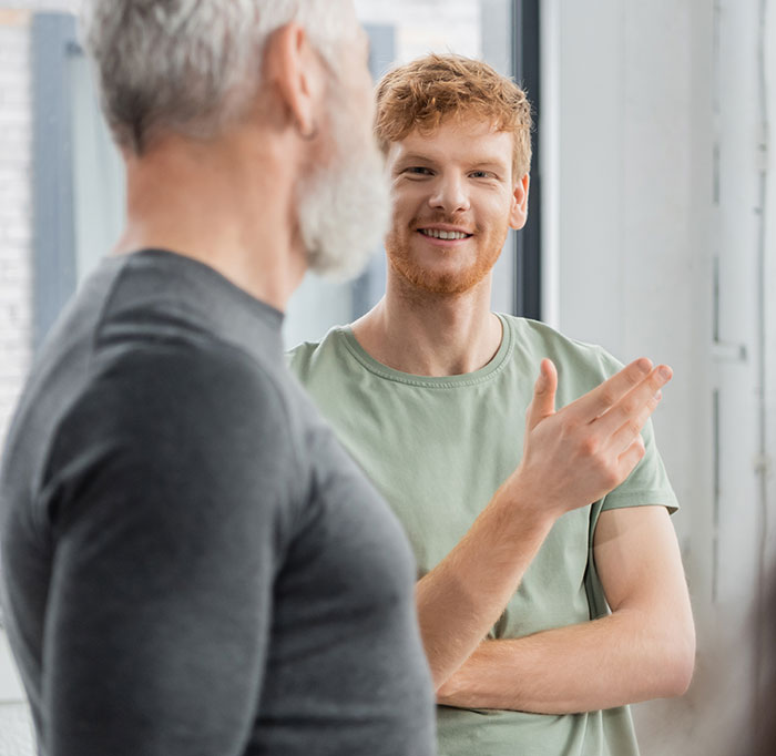 Young man smiling and gesturing while discussing social cues with an older man in a bright indoor setting.