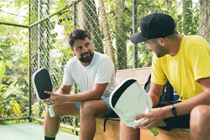 Two men with high social skills chatting and holding paddles, sitting on a bench near a chain-link fence outdoors.