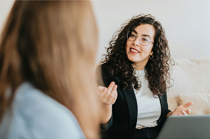 A woman with glasses and curly hair demonstrating high social skills while engaging in a friendly conversation indoors.