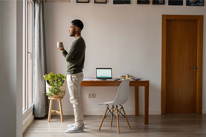 Man with high social skills standing by window with coffee cup in modern room, laptop on wooden desk nearby.