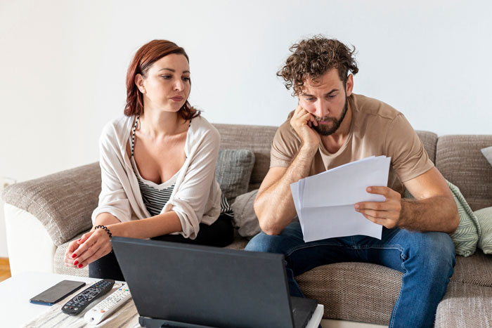Young man mooching off parents looks worried while woman watches, both sitting on couch with laptop and papers.