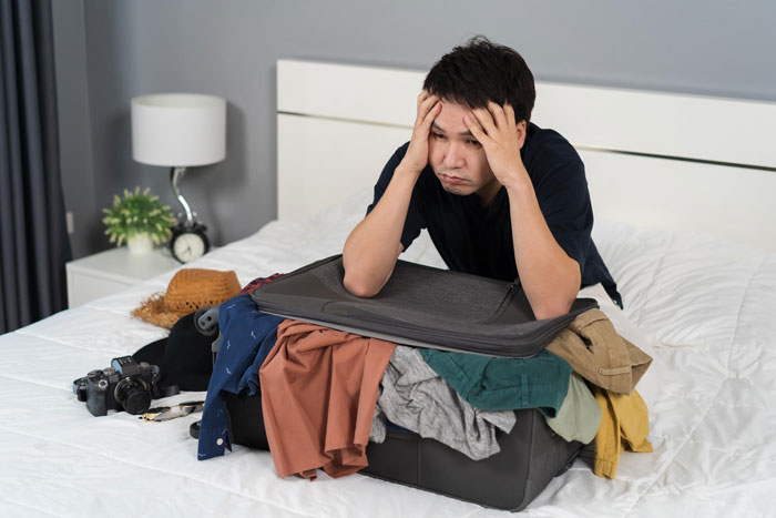 Young man looks stressed sitting on bed with overstuffed suitcase after parents kick him out for stealing money.