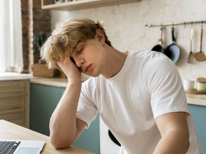 Young man in a white shirt looking stressed at a kitchen table, reflecting on career challenges against dreams.