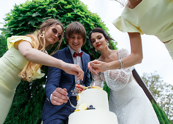 Step-dad looking sad and distant while others around him happily cut a wedding cake outdoors in a garden setting.