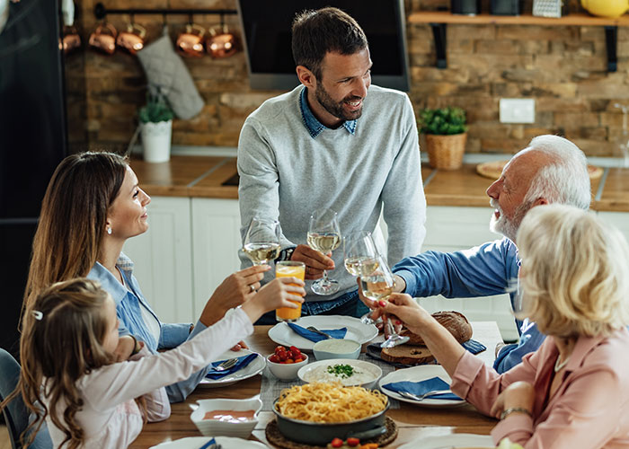 Step-dad sharing emotional speech while family gathers around kitchen table raising glasses in somber moment.