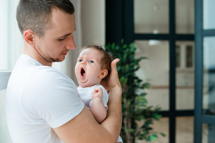A father holding a yawning baby indoors, symbolizing family dynamics affected by mom poisoning kids against stepmom.