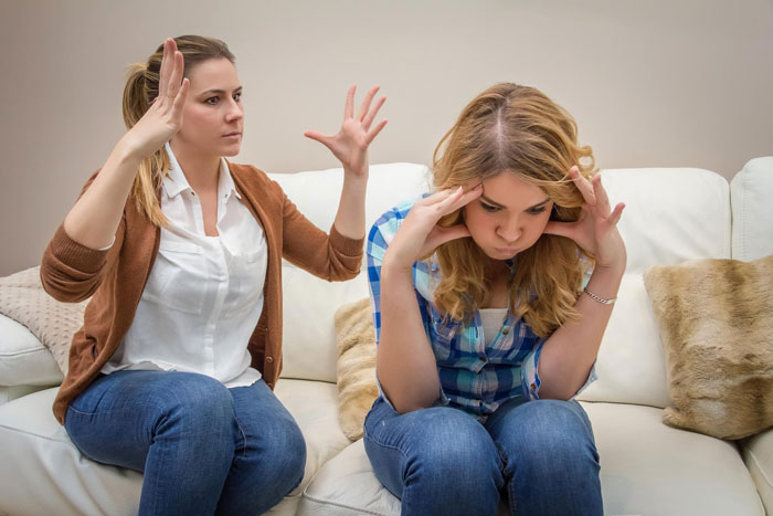 Stepmother raising hands in frustration while controlling family relationship with upset teenage girl on couch Stepmother raising hands in frustration while controlling family relationship with upset teenage girl on couch