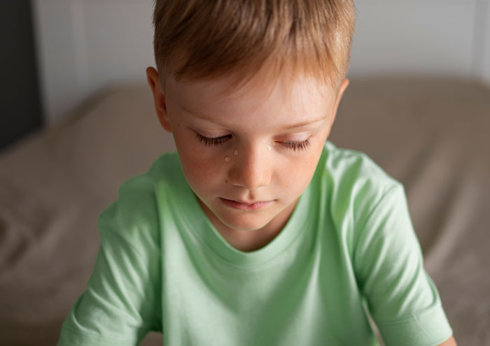 Young boy with a tear on his cheek, sitting on a bed, illustrating family room size debate involving biological daughter.