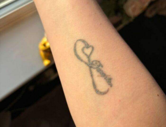Close-up of a small black ink tattoo on a forearm, showing a heart and infinity symbol design refused by tattoo artists.