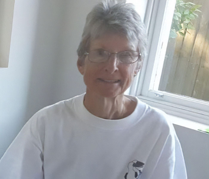 Elderly woman wearing glasses and white shirt sitting indoors near a window, representing stranded on a remote island.