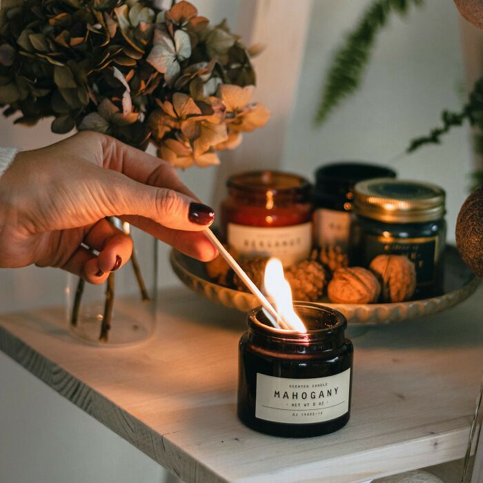 Hand lighting a mahogany scented candle with dried flowers and nuts in the background, cozy home setting disasters playdates.