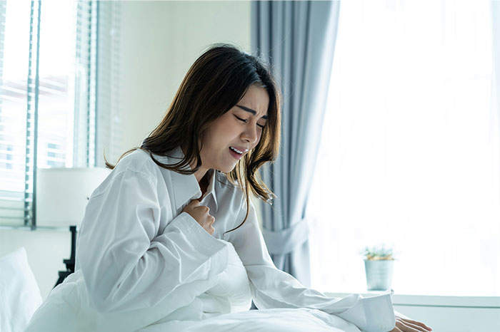 Young woman in white shirt sitting on bed clutching chest in pain depicting symptoms that made people see a doctor for cancer diagnosis