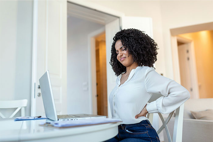 Woman holding her back in pain while sitting at a table with a laptop, illustrating symptoms that led to cancer diagnosis.