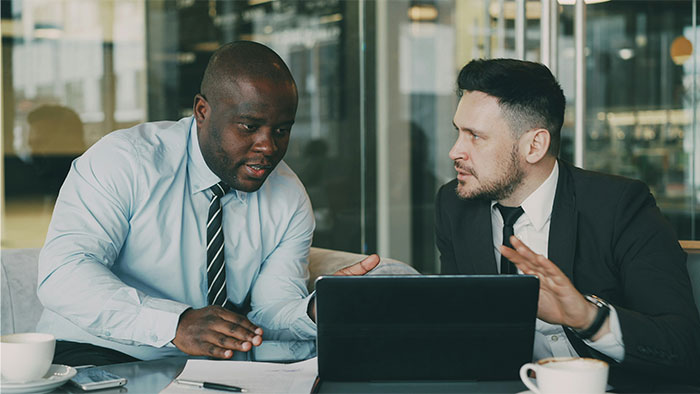 Two men in business attire discussing symptoms that made people see a doctor and it turned out to be cancer using a tablet.