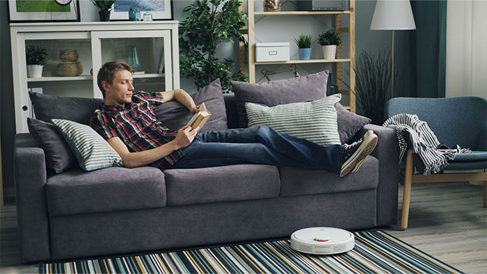 Man relaxing on a couch reading a book in a living room with a robot vacuum, related to symptoms that led to cancer diagnosis.