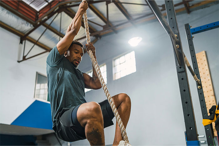 Man climbing rope in gym, focused on fitness, illustrating strength despite symptoms linked to cancer diagnosis.