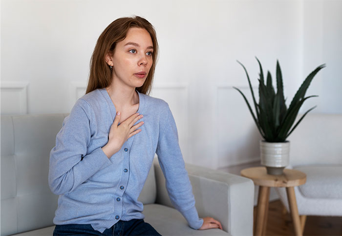 Young woman sitting on couch holding chest, appearing concerned about symptoms that made people see a doctor for cancer.