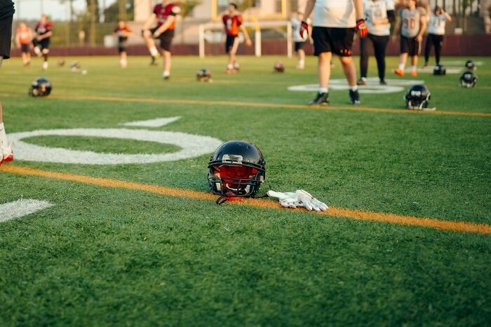 Football helmet and gloves on grass field with players practicing, symbolizing shocking and dark secrets in sports.