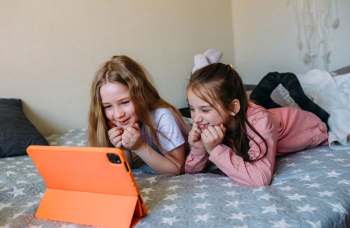 Two daughters lying on bed smiling while watching a tablet, representing taking daughters get nails done experience.