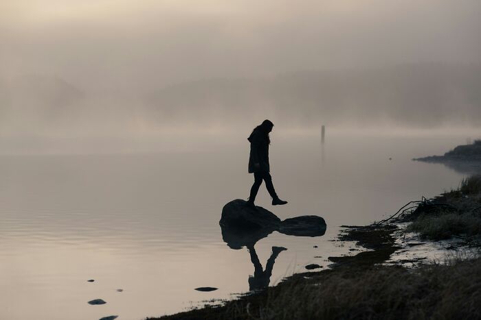 Silhouette of a person standing on rocks by foggy water, reflecting on dark secrets that could ruin lives if revealed.