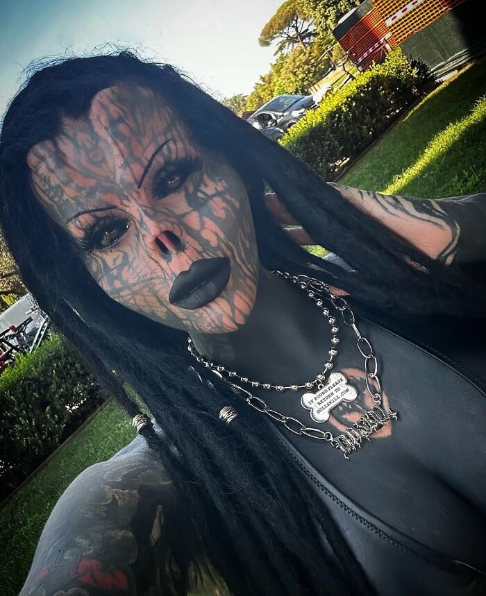 Tattooed woman with extensive body art and no nose, showcasing her unique appearance outdoors with dreadlocks and dark makeup.