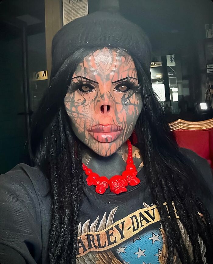 Tattooed woman with extensive facial tattoos and missing nose wearing red skull necklace and Harley-Davidson shirt indoors.