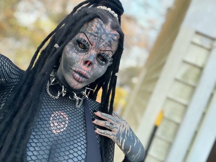 Tattooed woman with black facial tattoos and cut off nose posing outdoors wearing a fishnet top and spiked collar.