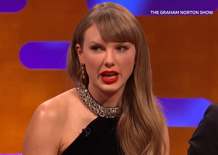 Taylor Swift speaking on the Graham Norton Show with styled hair and red lipstick in a black outfit.
