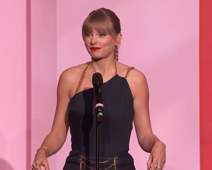 Taylor Swift speaking at a microphone during a live event, fans discussing her Graham Norton appearance and reactions.