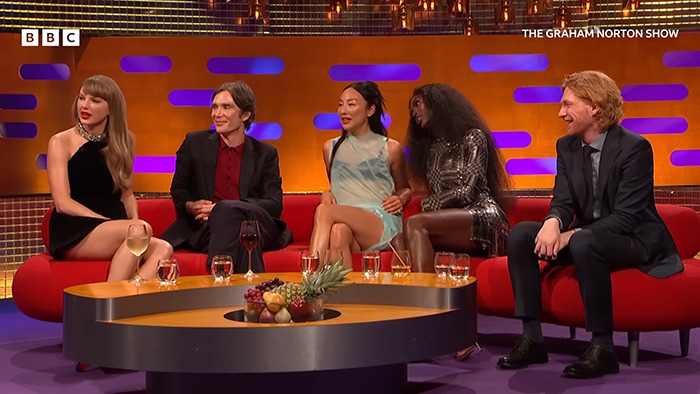 Taylor Swift and guests seated on a red couch during Graham Norton Show appearance with drinks on table in front.