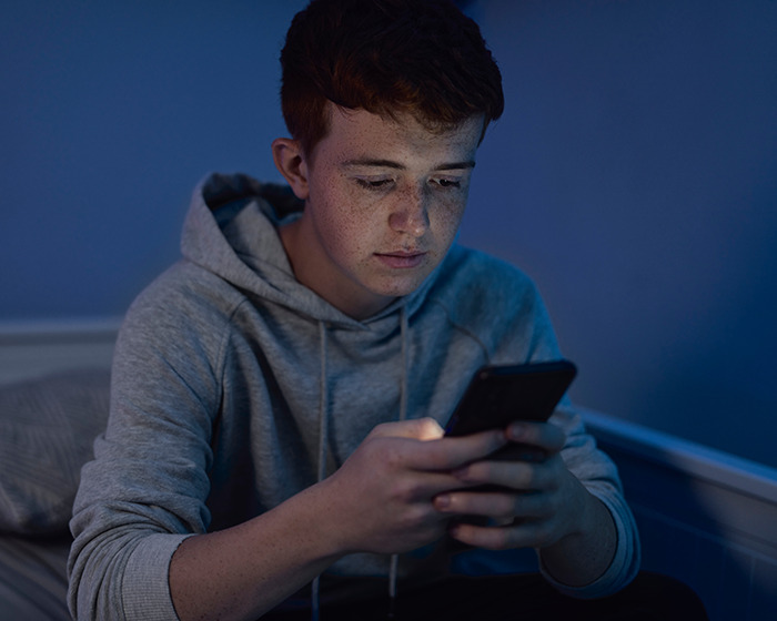 Teen with freckled face wearing gray hoodie, looking at smartphone in dimly lit room, illustrating family dinner leads to arrest story.