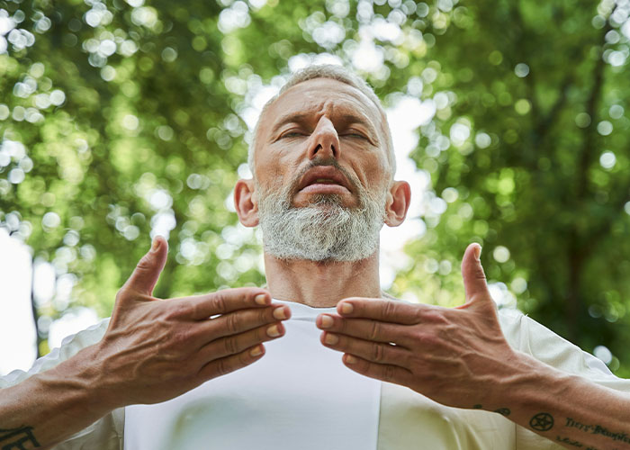 Middle-aged man with gray beard practicing breathing exercise outdoors amid lush green trees, demonstrating unusual breathing method. Middle-aged man with gray beard practicing breathing exercise outdoors amid lush green trees, demonstrating unusual breathing method.