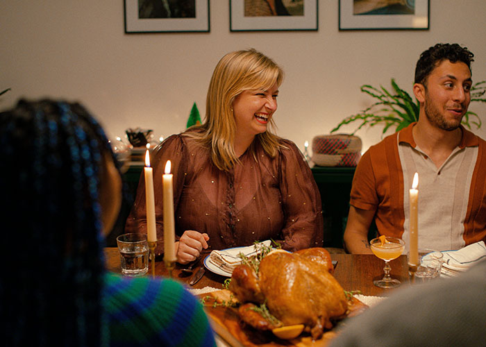 Group gathered around a Thanksgiving dinner table with a roasted turkey, candles, and people socializing and smiling.