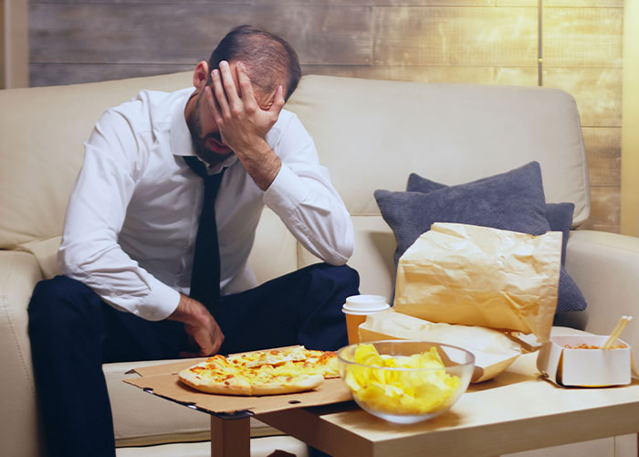 Stressed man sitting on couch with hand on face next to leftover pizza and snacks, reflecting Thanksgiving misery and divorce feelings.
