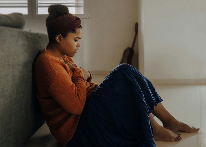 Woman in an orange sweater and blue skirt sitting on floor, looking upset on Thanksgiving during a difficult divorce time.