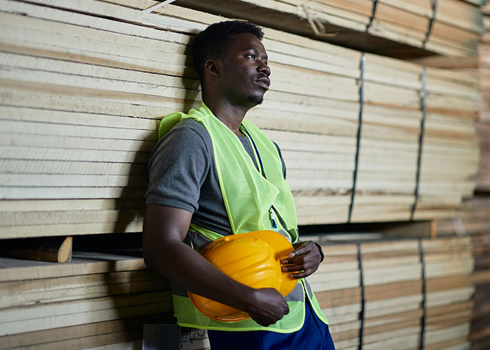 Man in safety vest holding yellow hard hat, looking pensive while leaning against stacked wooden boards in workshop.