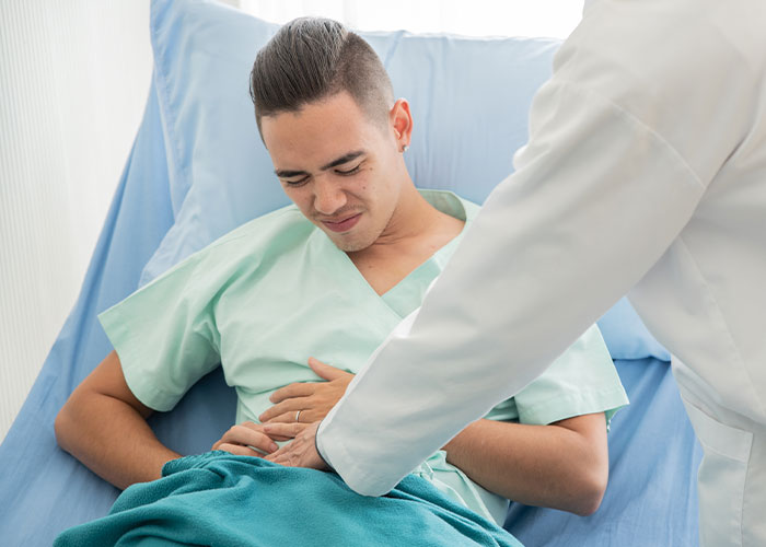 Young man in hospital gown looking uncomfortable while doctor examines his abdomen, illustrating misery on Thanksgiving.