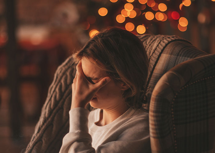 Woman sitting on a chair with hand covering face, looking stressed and miserable during the Thanksgiving holiday season.
