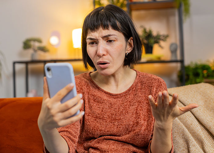 Woman in a cozy living room looking upset while holding a smartphone, expressing frustration during Thanksgiving gatherings.