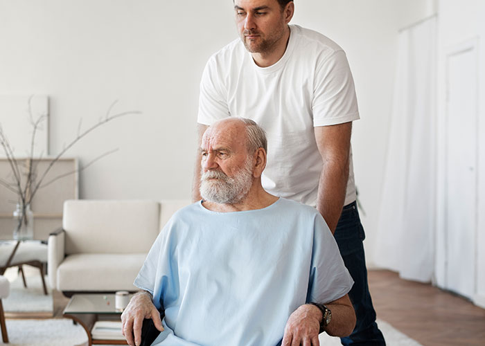 Elderly man in wheelchair looking upset while younger man stands behind him in a bright living room setting.