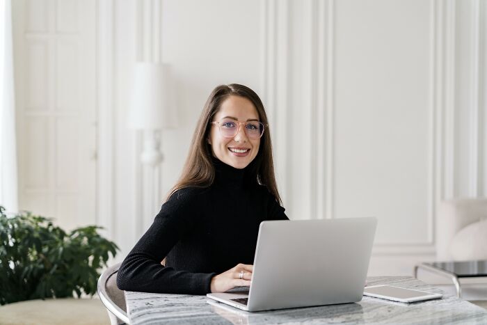 Young woman with glasses smiling, working on a laptop in a bright room about people who lost jobs to AI.