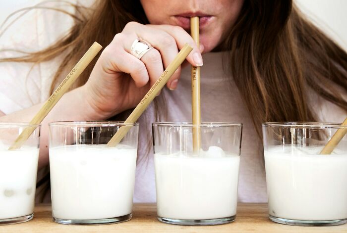 Person sipping a drink through a straw with four glasses of milk or a white beverage lined up on a wooden table.