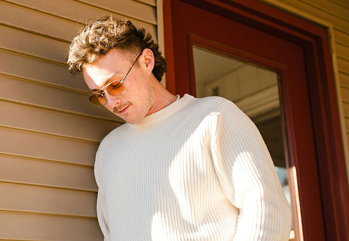 Young man wearing sunglasses and a cream sweater, standing outside near a house, seemingly unaware of what he’s seeing.