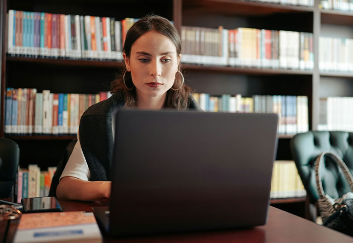 Young woman using laptop in library, focused on screen while realizing something she isn’t supposed to see.