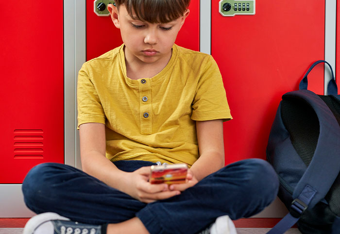 Young boy sitting on floor by lockers, looking surprised as he sees something unexpected on his phone screen.