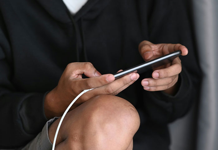 Person in black hoodie holding a smartphone with charging cable, capturing moments they aren’t supposed to see.