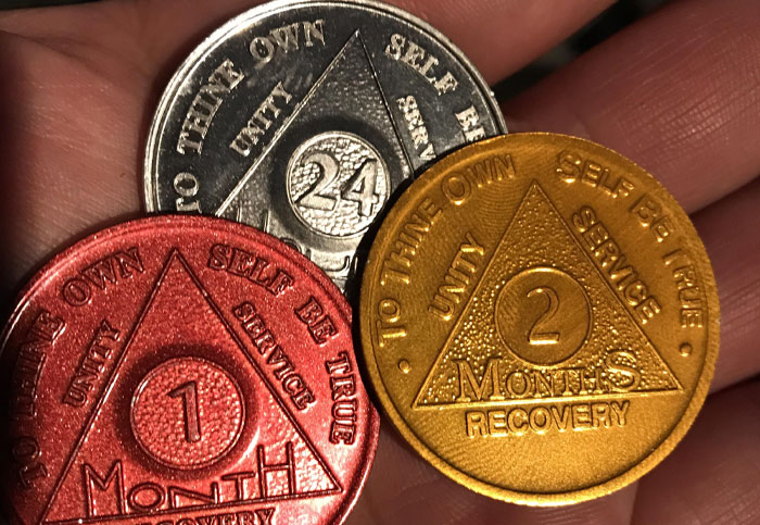 Close-up of recovery coins held in a hand, showing symbols of unity, service, and self-recovery milestones.