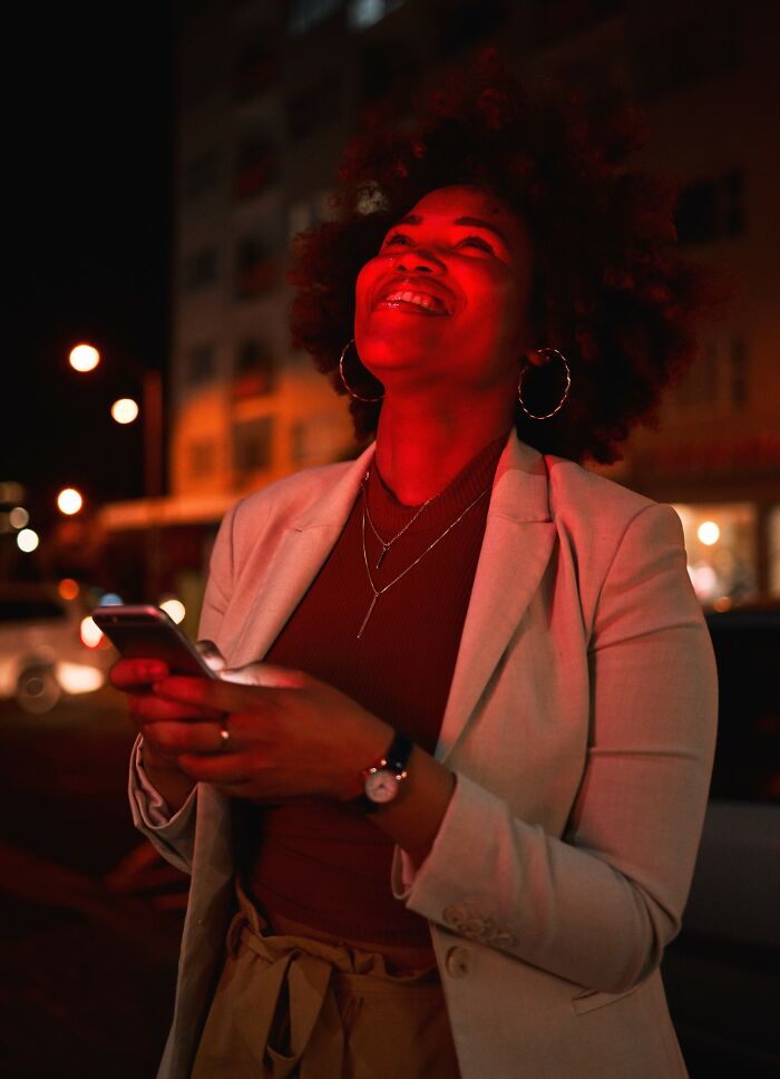 Young woman smiling and holding phone on a city street at night, illustrating drunk fumbles and funny moments outdoors.